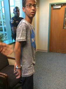 Ahmen Mohamed, a 14 year old brown skinned boy in a grey shirt and jeans, stands with his hands cuffed behind his back with a police officer in the background.